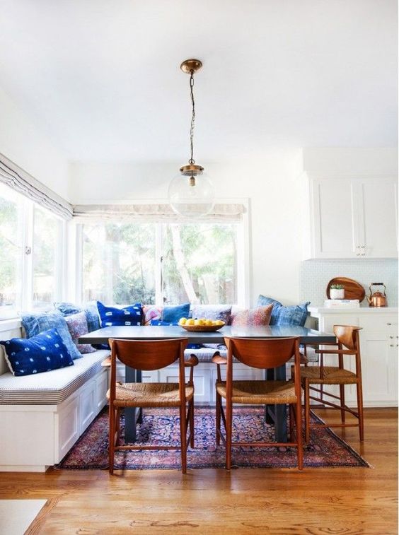 a boho dining nook with a corner window, a large upholstered banquette seating, a dining table and woven chairs