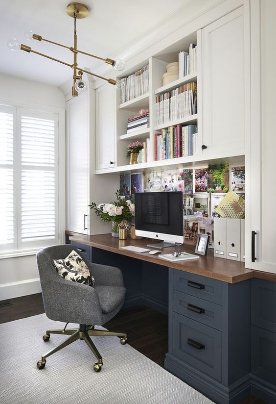 a modern farmhouse home office with two tone kitchen cabinets, a desk with a butcherblock countertop, a grey chair and a vintage chandelier