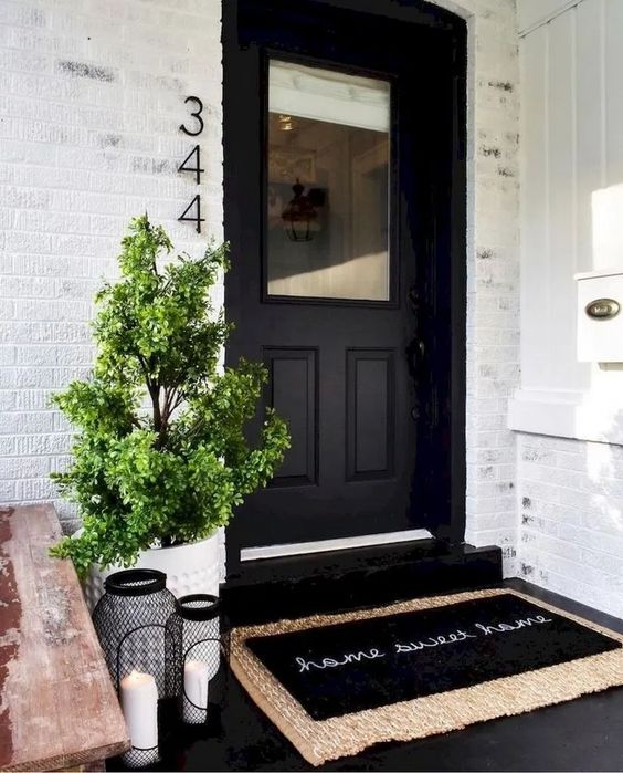 a laconic modern porch with a black glass door, layered rugs, black candle lanterns and a planter with a mini trees is a cool space