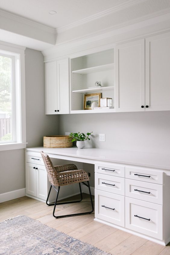 a modern neutral home office with white shaker style cabinets, a desk with lots of storage, a rattan chair and some plants