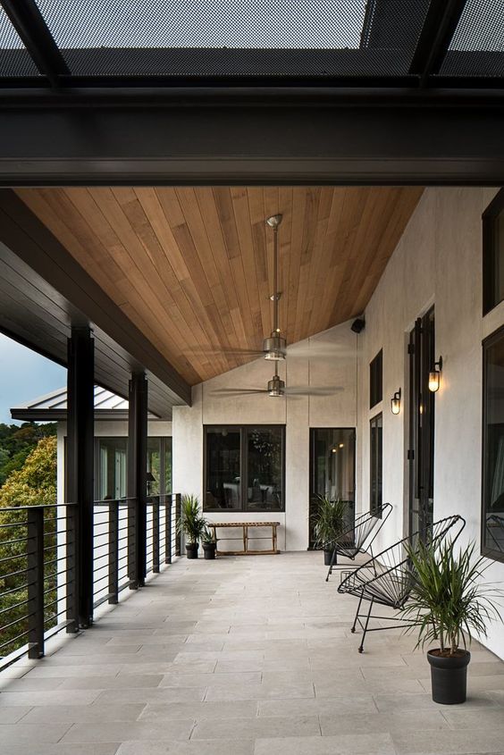 a modern porch clad with neutral tiles, with black woven chairs, potted plants, a bench and some more plants