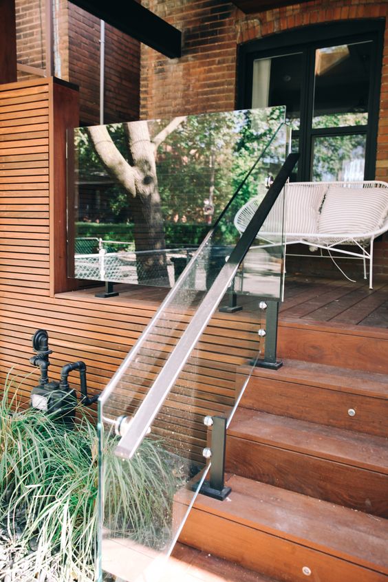 a modern porch clad with rich stained wood, with glass railings, grasses around and a white metal bench with pillows is chic