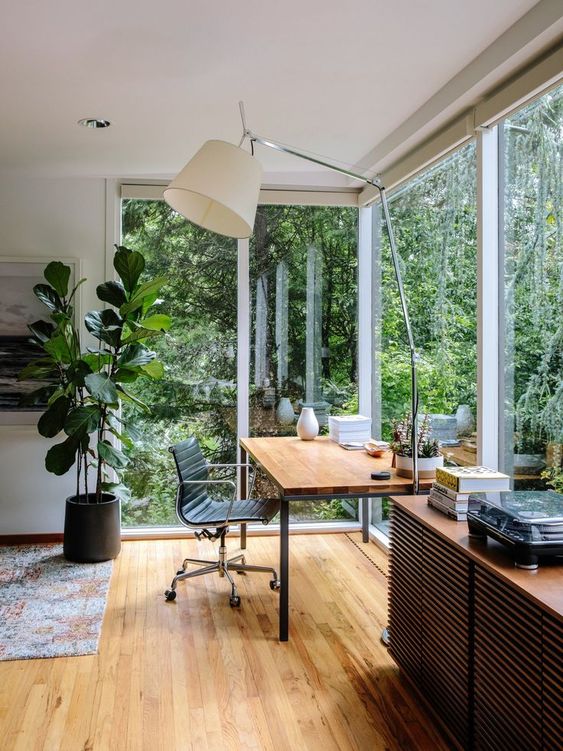 a beautiful and dreamy working space with a corner window, a desk, a black leather chair, a calming forest view and a large floor lamp