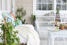 a beautiful farmhouse patio with a wodoen sofa and white rattan chairs, a vintage table, potted plants, colorful bedding and pillows