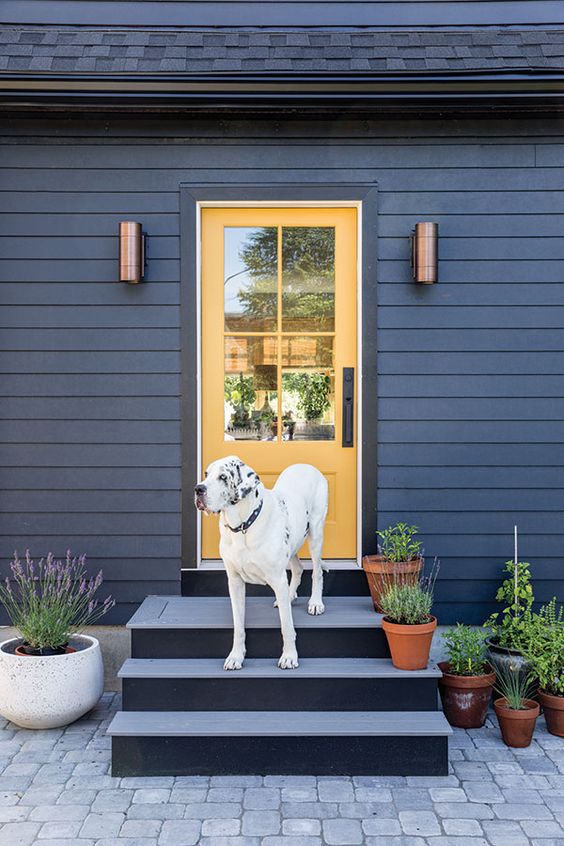 a bold Scandi front porch with a grey ladder, a yellow door, potted greenery and blooms and copper sconces