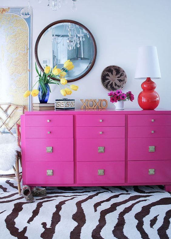 a bold space with a hot pink knob dresser, a zebra rug, a chic chandelier, a round mirror and a table lamp with a red base