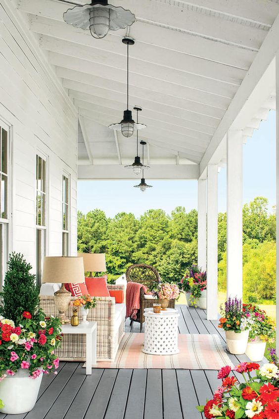 a bright and cool farmhouse porch with a white wicker sofa, potted blooms and plants, side tables and pendant and table lamps