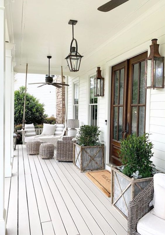 a charming farmhouse porch with a hanging bench with striped pillows, a wicker chair and poufs, plants in wooden boxes and rustic vintage lanterns