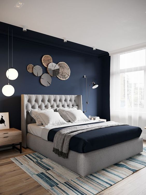 a chic bedroom with a navy accent wall, a grey upholstered bed, navy and grey bedding, a wood slice decoration and pendant lamps