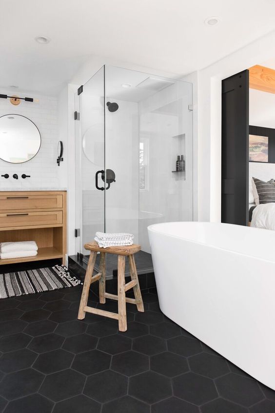 a chic fancy bathroom with a black hex tile floor, an oval tub, wooden furniture and a striped rug plus a glass enclosed shower