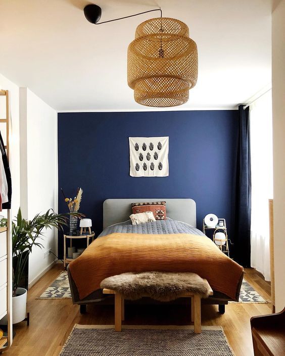 a chic farmhouse bedroom with a navy accent wall, a grey upholstered bed, pretty bedding, a woven pendant lamp and some potted plants
