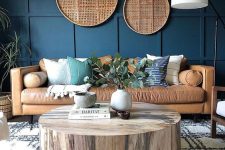 a chic farmhouse living room with a navy paneled accent wall, a tan leather sofa, burlap decor, a tree stump table and greenery