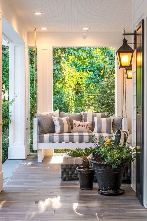 a cool farmhouse porch with a striped upholstered sofa, a wicker lounger, potted plants and greenery and lanterns on the walls