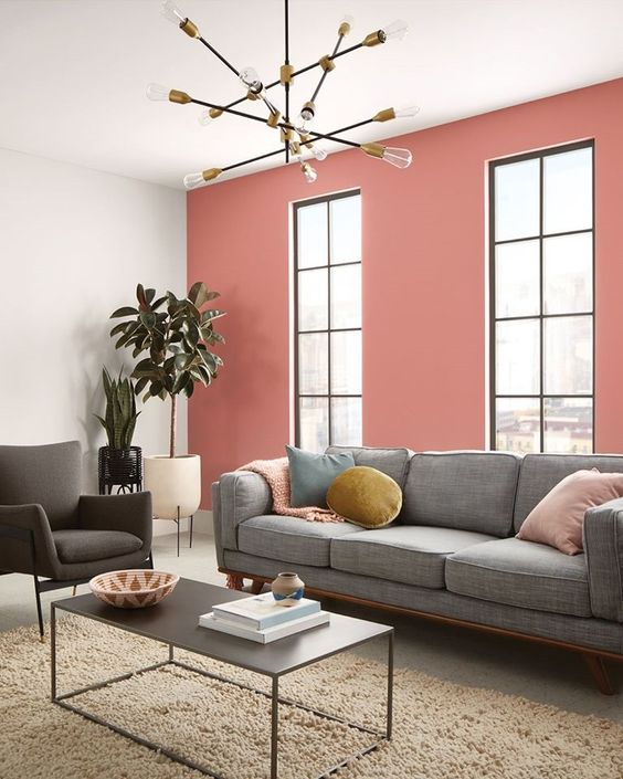 a cool modern living room with a pink accent wall, a grey sofa and colorful pilows, a black coffee table and a catchy chandelier