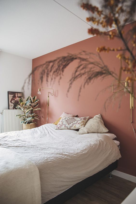 a dreamy and relaxed bedroom with a dusty pink accent wall, a bed and printed pillows, some potted plants and gilded sconces