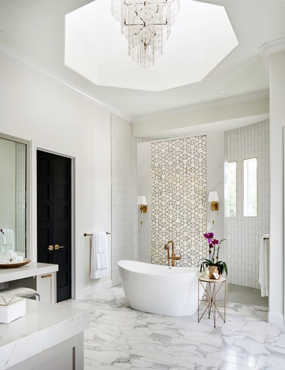 a fabulous fancy bathroom with white tiles and patterned ones, white marble tiles on the floor, a crystal chandelier and brass touches