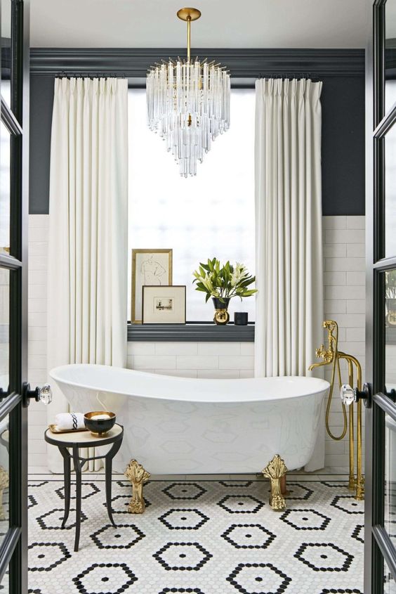 a luxorious bathroom with a black and white tile floor, white subway tiles and black walls, a chic tub, gold fixtures and a lovely chandelier