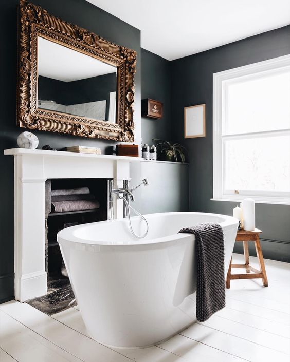 a fancy bathroom with black walls and a white planked floor, a fireplace used for storign towels, an oval tub and a mirror in a refined frame