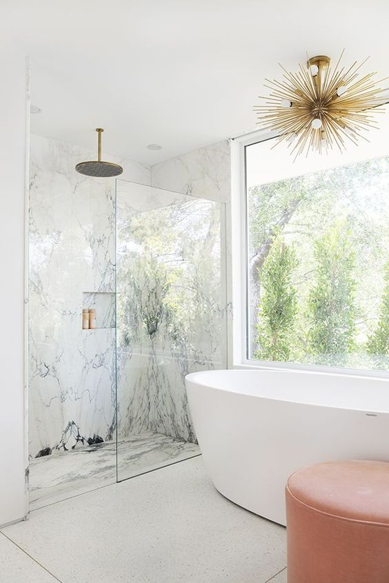 a fancy bathroom with white marble in the shower space, a glazed wall, a bathtub, a coral ottoman and a gold sunburst chandelier
