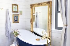 a fancy bathroom with white planked walls, a dark stained floor, a vintage navy bathtub, a floor mirror in a gilded frame, a mini gallery wall and a crystal pendant lamp