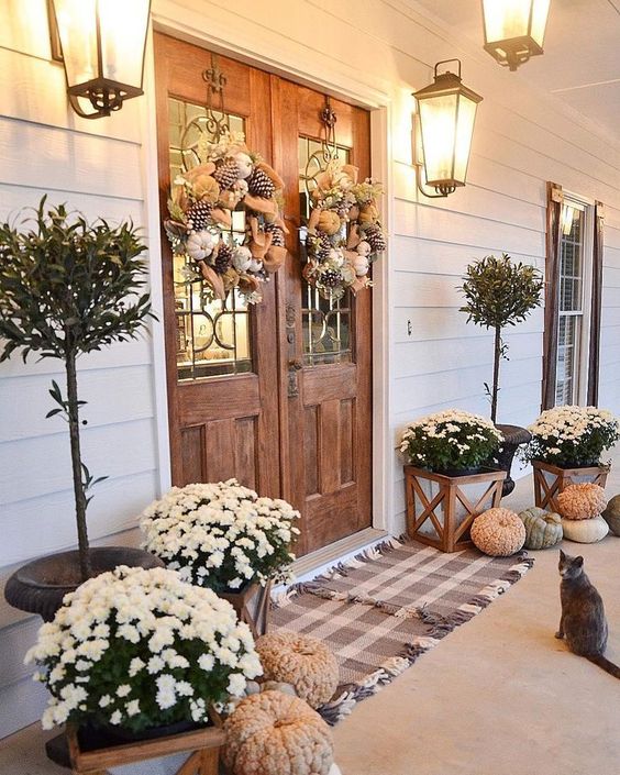 a farmhouse fall porch with plaid rugs, potted blooms, heriloom pumpkins, potted trees and feather wreaths plus wall lanterns
