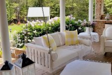 a farmhouse porch with a white upohlstered bench on chains, several candle lanterns, a white wicker chair and some pretty textiles to cozy up the space