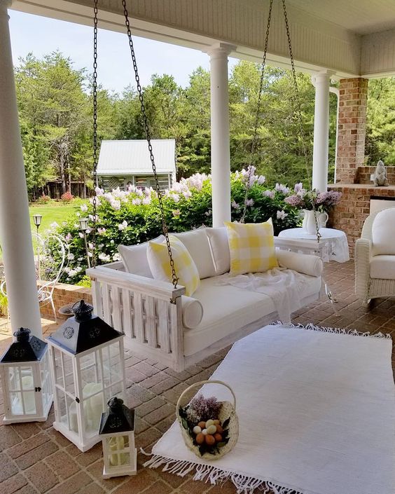 a farmhouse porch with a white upohlstered bench on chains, several candle lanterns, a white wicker chair and some pretty textiles to cozy up the space