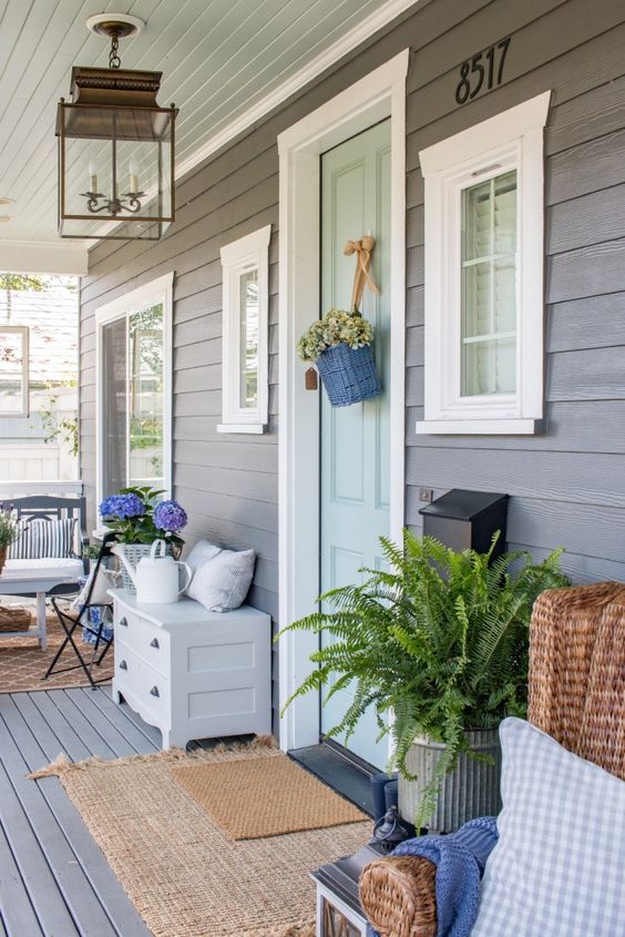 a farmhouse porch with a wicker chair and printed textiles, a suspended bench, layered rugs, a sideboard and a watering can plus blooms and greenery