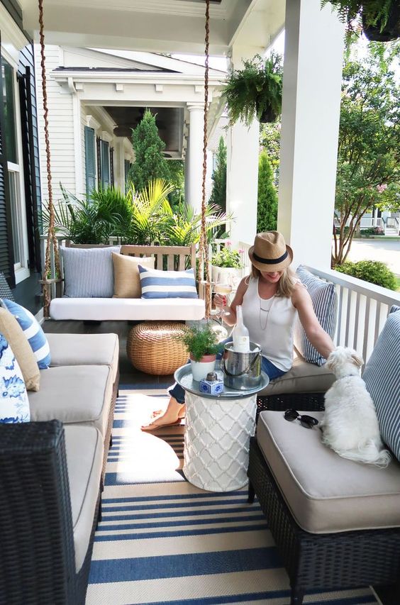 a farmhouse porch with black wicker furniture, a hanging upholstered bench, side tables and poufs and a striped rug plus pillows