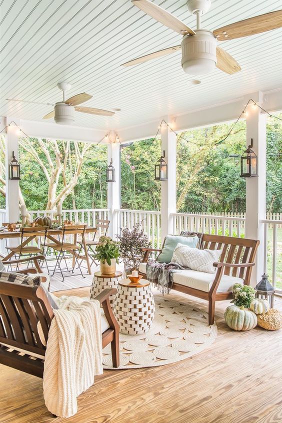 a large and welcoming farmhouse porch with a simple wooden dining set, wooden benches with upholstery, side tables and potted plants