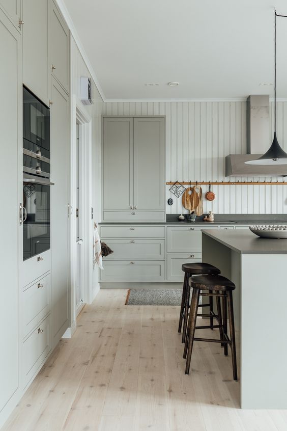 a light grey modern country kitchen with shaker style cabinetry, black countertops and built-in appliances, a modern hood and tall stools