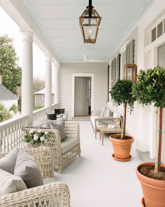 a lovely modern farmhouse porch with neutral wicker furniture, an upholstered bench and chairs, potted trees and lanterns