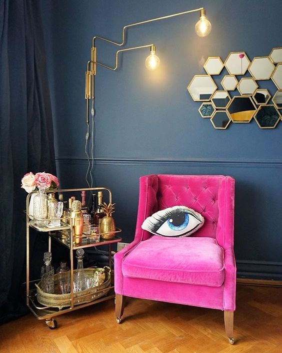 a lovely nook with a navy wall and black curtains, a hot pink chair, a gold bar cart, a cool gold sconce and hexagon mirrors