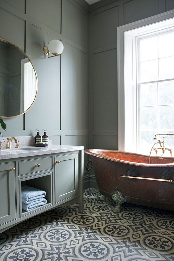 a modern country bathroom with grey green paneled walls and a matching vanity, a copper vintage bathtub and a lovely mosaic tile floor