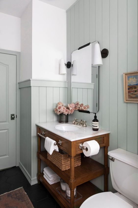 a modern country bathroom with pale aqua paneling, a stained vanity, white appliances, vintage lamps and some blooms