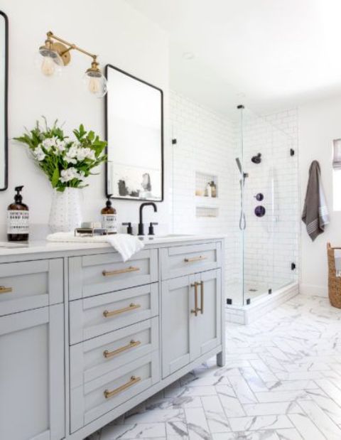 a modern country bathroom with white marble herringbone tiles, white subway ones, a light grey vanity and a shower space plus brass touches