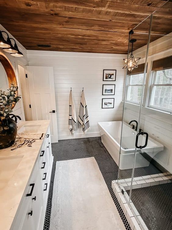 a modern country bathroom with white planked walls and a stained ceiling, a penny tile floor, white furniture and appliances and black fixtures