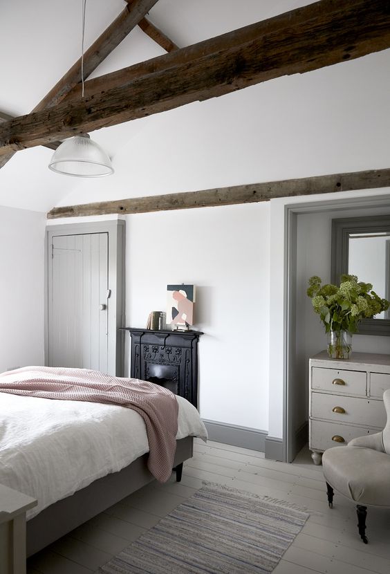 a modern country bedroom with dark stained wodoen beams, a non-workign fireplace, neutral furniture and neutral textiles