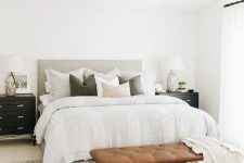 a modern country bedroom with neutral walls, a grey upholstered bed, a leather bench, black dresser nightstands and neutral textiles