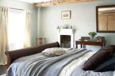 a modern country bedroom with wooden beams, a fireplace, a dark stained bed and a mirror plus some vintage furniture