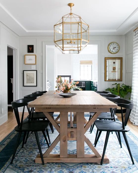 a modern country dining space with a stained trestle dining table, black chairs, a blue rug and a gilded faceted chandelier
