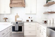 a modern country kitchen with shaker style cabinets, a white subway tile backsplash, a wooden hood and floating shelves