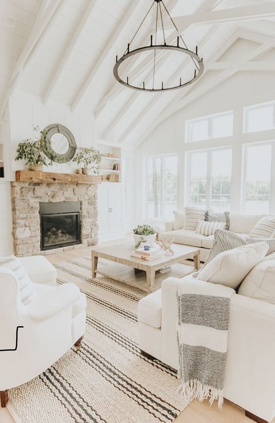 a modern country living room designed in neutrals, with a fireplace clad with stone, cozy furniture and printed textiles