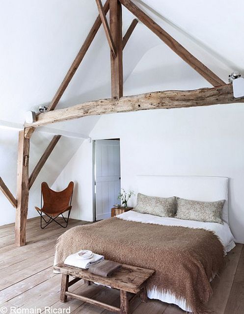 a modern farmhouse bedroom with stained wooden beams that highlight the attic ceiling, a neutral bed with a bench and a leather chair