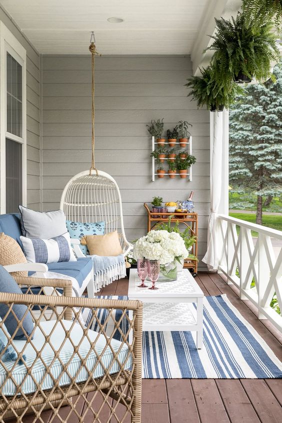 a modern farmhouse coastal porch with wicker furniture, a suspended egg shaped chair, a coffee table, striped accessories, potted plants