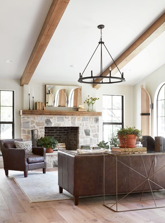 a modern farmhouse living room with a stone clad fireplace, wooden beams, leather furniture, a metal chandelier and potted plants