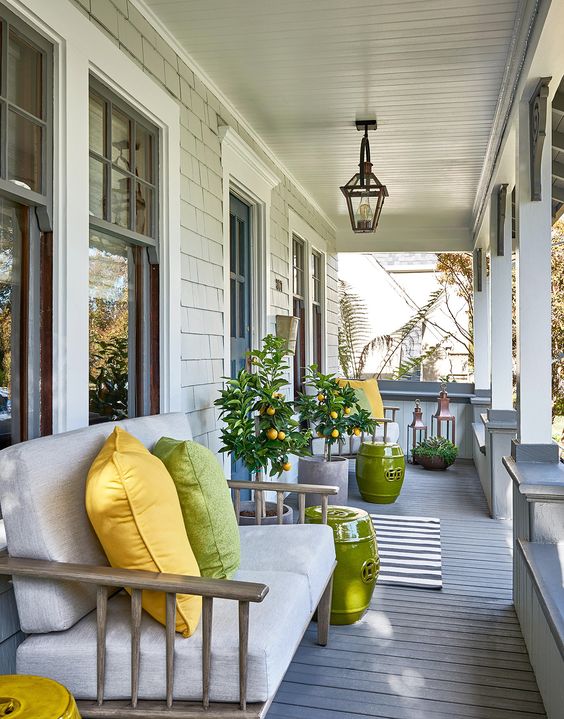 a modern farmhouse porch styled for summer, with simple wooden furniture, bright pillows, bold green side tables and potted trees