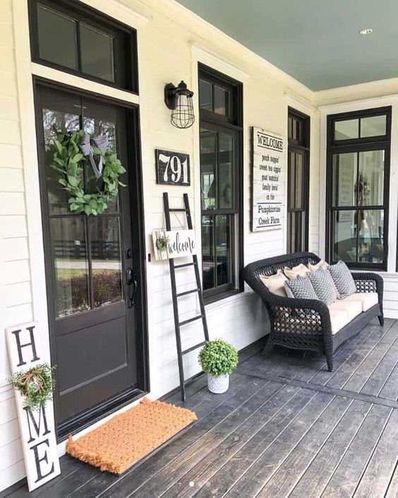 a modern farmhouse porch with a black wicker sofa with lots of pillows, a ladder, some signs and a greenery wreath