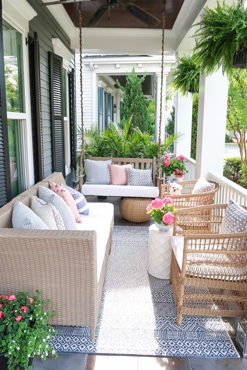 a modern farmhouse porch with wicker furniture, printed pillows, a suspended bench, potted blooms and greenery is welcoming