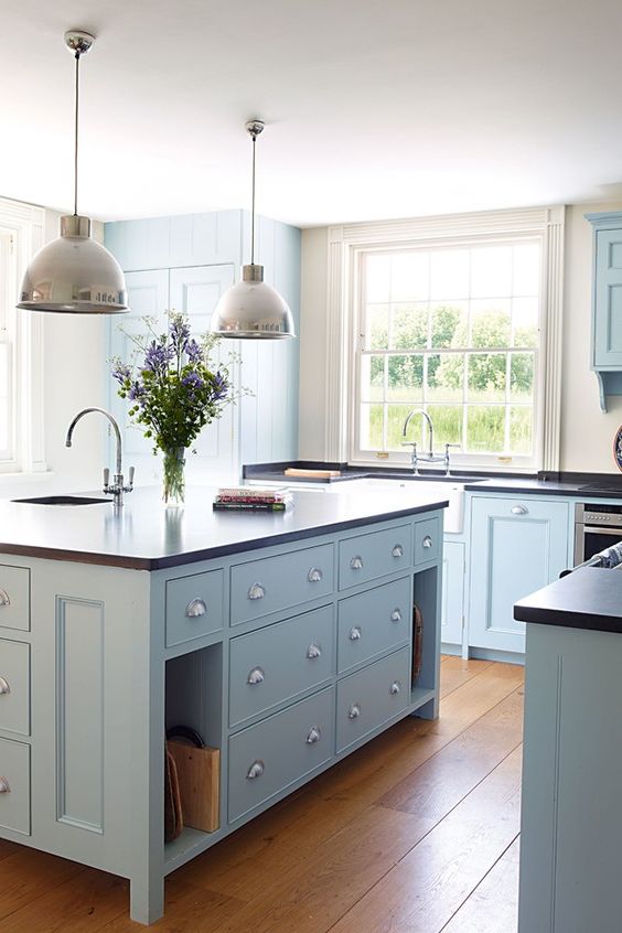 a modern light blue country kitchen with dark countertops, pendant lamps and large windows plus a lovely hardwood floor is chic